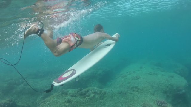 SLOW MOTION, UNDERWATER: Athletic man on epic vacation dives out of the turquoise ocean on his surfboard. Extreme male surfboarder does a duck dive on his way to catch another epic Cloudbreak wave.