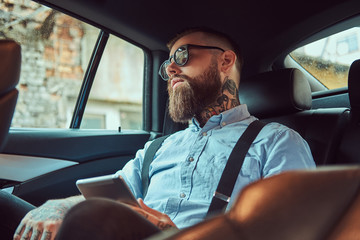 Old-fashioned tattooed hipster guy in a shirt with suspenders sitting in a luxury car on back seat.