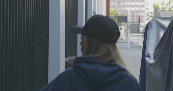 Back view of trendy girl wearing cap and blue sweatshirt walks down alley.. City urban style. 4K video shooting by handheld gimbal