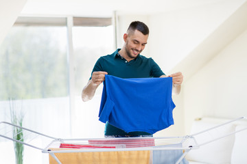 laundry and household concept - happy man taking clothes from drying rack and putting them to basket at home