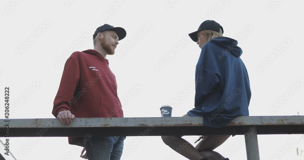 Poster Bottom view of two people talking to each other. Beautiful young couple of man and woman standing on the edge of the building roof. City urban style. 4K video shooting by handheld gimbal