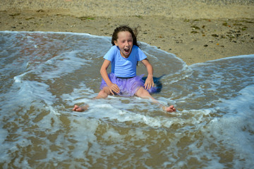 Cheerful little girl sitting on the yellow sand on the beach on a Sunny day in a purple dress, playing and laughing when the wave rolls on her. Horizontal orientation