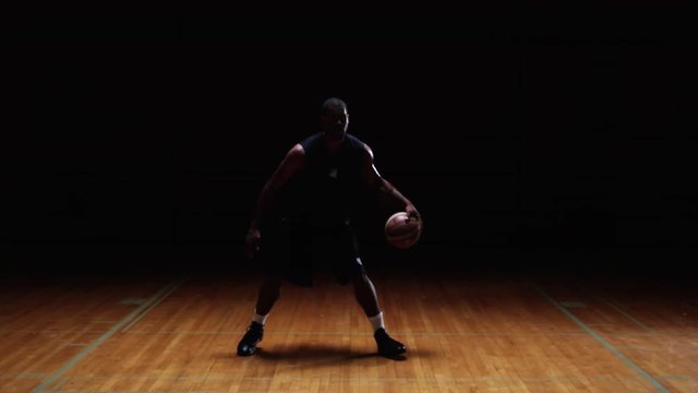 A basketball player dribbles the ball through his legs on a dark court