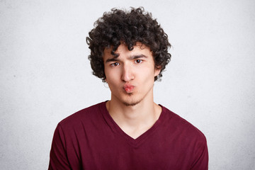 Obraz premium Attractive young male with crisp dark hair keeps mouth round, makes grimace, wears casual sweater, looks directly into camera, isolated over white studio concrete wall. People, youth concept