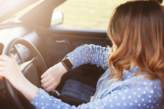 Photo Of Attractive Female Checks Time On Watch, Sits In Car, Stuck In Traffic Jam And Being Late For Meeting With Colleagues, Has To Delay It. Lovely Woman Rides Auto . People, Driving And Hurry