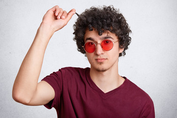 Attractive bearded male shows his dark crisp hair, has specific appearance, wears trendy red shades and t shirt, poses against white background. Fashionable hipster guy poses in studio. People, style