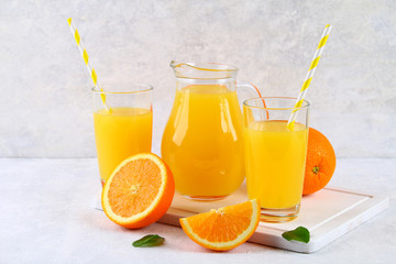Glass cups and a pitcher of fresh orange juice with slices of orange and yellow tubes on a light gray table.
