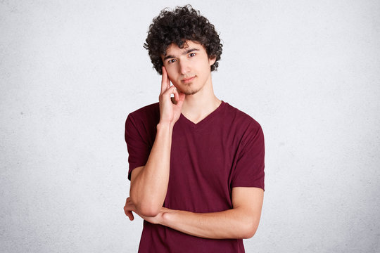 Waist up portrait of handsome male keeps hand near temple, has pensive and satified expression, wears casual clothing, poses against white background. People, attitude and facial expressions