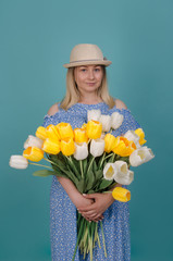 Woman with flowers in her hands. Young beautiful woman in straw hat wearing blue dress with white and yellow tulips. Summer and spring concept. Isolated on blue studio background