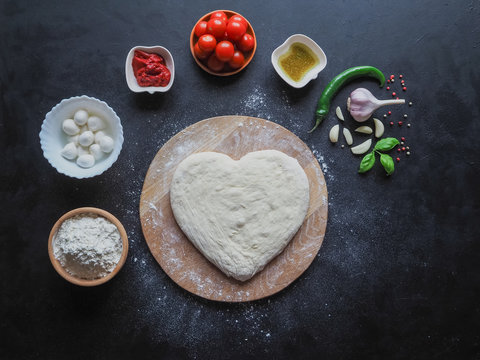 Heart-shaped dough and a set of ingredients for pizza on a black table. The view from the top.