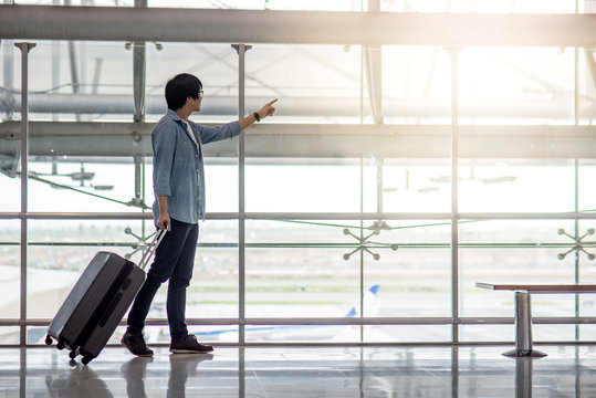 Young Asian man walking with suitcase luggage in the airport terminal pointing the plane taking off, travel lifestyle