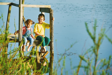 Brother and sister sitting on the bridge . Children have fun spending summer holidays at the pond