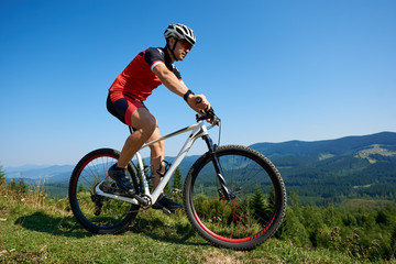 Fototapeta na wymiar Muscular tourist cyclist in helmet, sunglasses and full equipment riding bike on grassy hill. Mountains and blue summer sky on background. Active lifestyle and extreme sport concept