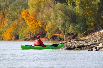 A girl in a green kayak and a red lifejacket is rowing along the Danube River against the...