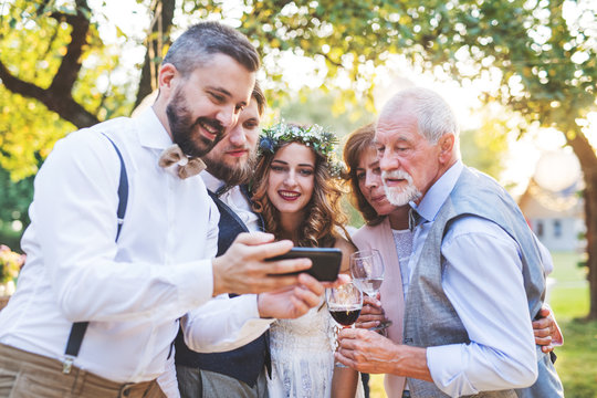 Bride, groom and guests with smartphones taking selfie outside at wedding reception.