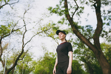 Portrait of young athletic beautiful brunette girl in black uniform and cap standing, resting and looking aside after running, training in city park outdoors. Fitness, healthy lifestyle concept.