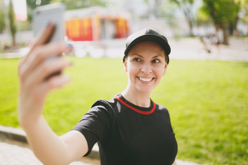Young athletic smiling beautiful brunette girl in black uniform, cap looking on smartphone and doing selfie on mobile phone during training in city park outdoors. Fitness, healthy lifestyle concept.