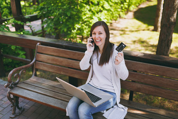 Female holding credit card, talking on mobile phone, having good pleasure conversation. Woman sitting on bench working on modern laptop in street outdoors. Mobile Office. Freelance business concept.