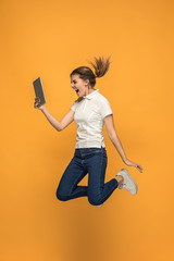 Image of young woman over orange background using laptop computer or tablet gadget while jumping.