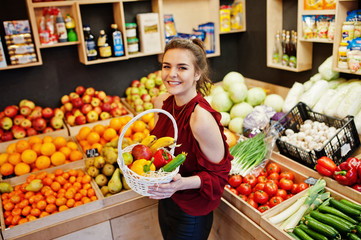 Girl in red holding different fruit and vegetables at basket on fruits store.
