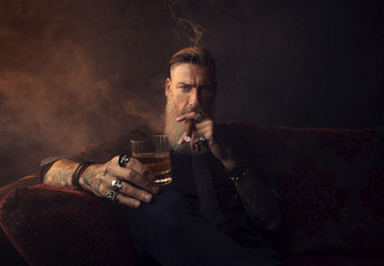 Mann mit Bart sitzt auf dem Couch, trinkt Whiskey und raucht eine Zigarre
