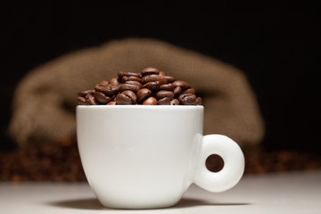 Eine weisse Kaffee-Tasse mit Kaffee-Bohnen und Kaffee-Sack auf dunklem, schwarzen Hintergrund