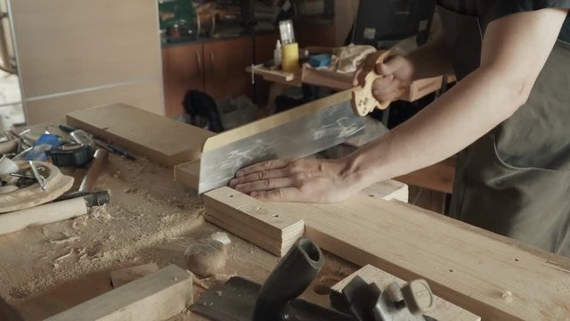 Skilled craftsman carpenter saws a workpiece of wooden board with a hacksaw in a workshop. Making handmade furniture.