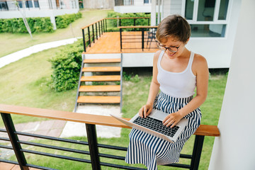 Attractive Young woman in summer dress with stripes and white tank top at the laptop keyboard on a green terrace background. Freelance and Internet work.