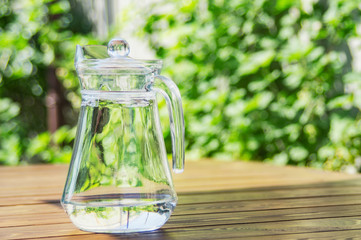 Pitcher with water on the wooden table against green foliage background.