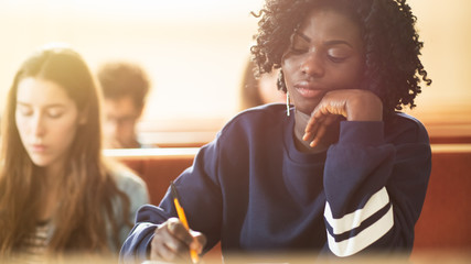 Tired Black Female Student Sitting Among Her Fellow Students in the Classroom, She's Writing in the Notebook and Listens to a Lecture.