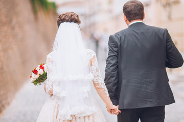 Happy newlywed couple on a walk in old European town street, gorgeous bride in white wedding dress...