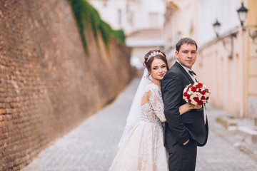 Happy newlywed couple on a walk in old European town street, gorgeous bride in white wedding dress...