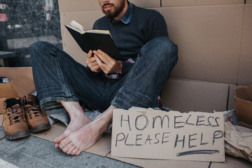 Smart homeless man is sitting on cardboard and reading a book. He is very concentrated on that. There is a sigh which says homeless please help.
