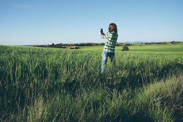 Naklejka na ściany i meble Mujer joven en una pradera verde sacando fotos con su teléfono móvil 