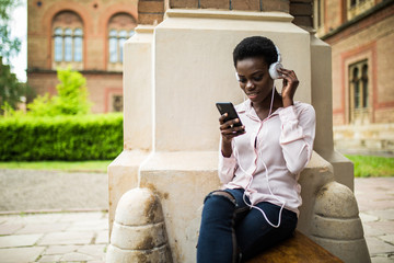 Beautiful afro american girl listening to music through headphones sitting on a bench. female with afro hair watching video on smartphone relaxing outside