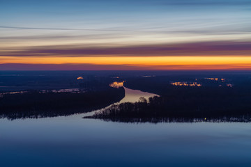 Beautiful river with big island with trees under predawn sky. Bright yellow stripe in picturesque cloudy sky. Early blue sky reflected in water. Colorful morning atmospheric image of majestic nature.