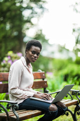 Work at break. Young African-American female with the laptop is sitting on the wooden bench. cheerful cute black girl on the park bench using the netbook.