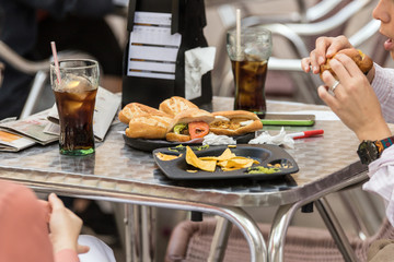 Girls sitting at a table eating a snack and drinking soda from cola outdoors. Friends eating street food sandwiches and fries.
