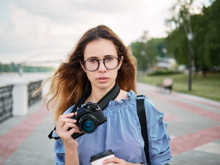 Young beautiful student girl in blue blouse and trendy glasses having fun with cup of coffee and photo camera posing on a river pier in city park
