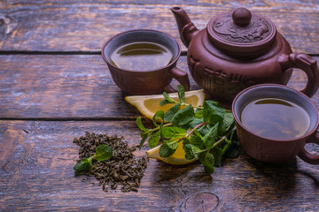 Green tea in a brown pot, cups with tea, mint, lemon, ginger on a dark old background.