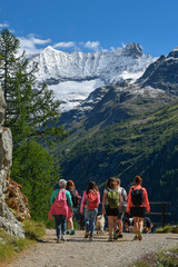 Fototapeta na wymiar Group of people with backpacks walking on a mountain path along a turquoise lake with snowy peaks on background. A beautiful sunny day to trekking with family and friends climbing to the snow-covered 