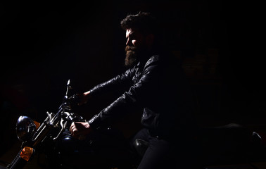 Night rider concept. Hipster, brutal biker in leather jacket riding motorcycle at night time, copy space. Man with beard, biker in leather jacket sitting on motor bike in darkness, black background.