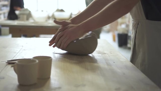 Young potter is kneading clay mass on table in pottery workshop. Man professional is in working process while standing at desk indoors. Guy kneads viscous mass on wooden surface, pressing it with