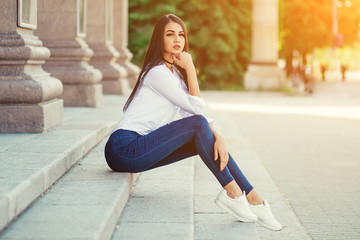 fashionable beautiful girl sitting outside in summer, brunette girl in jeans and white shirt, urban style
