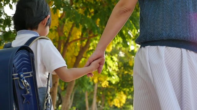Asian mother holding hand of little son with backpack outdoors, back to school slow motion 