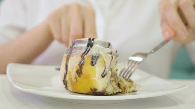 A woman in a white shirt eats with a knife and fork a bun cinnamon roll in a cafe and drinks coffee. Breakfast concept. 4k