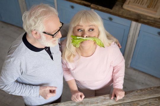 Adorable senior couple in kitchen. Sweet old man looking at his wife with love while she is being childish putting lettuce where mustache usually is.