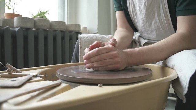 Portrait of male ceramist, who is woking in his bright studio. Man in dickey is sitting at throwing wheel with a piece of white clay on it, pouring water from sponge to the top and hangling the loam