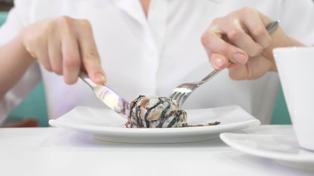 A woman in a white shirt eats with a knife and fork a bun cinnamon roll in a cafe and drinks coffee. Breakfast concept. 4k