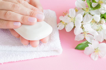 Obraz na płótnie Canvas White natural herbal soap in young, perfect, groomed woman's hands on the towel. Care about clean, soft and smooth skin. Beautiful branches of apple blossoms on pastel pink background. Fresh flowers.